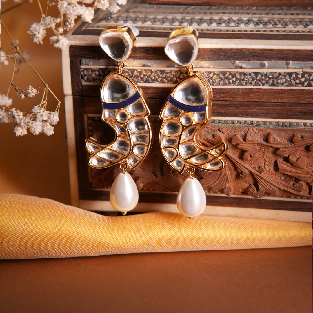 Ethnic Veera earrings with reflective stones, blue enamel, and hanging pearl drop on artistic vintage setting.