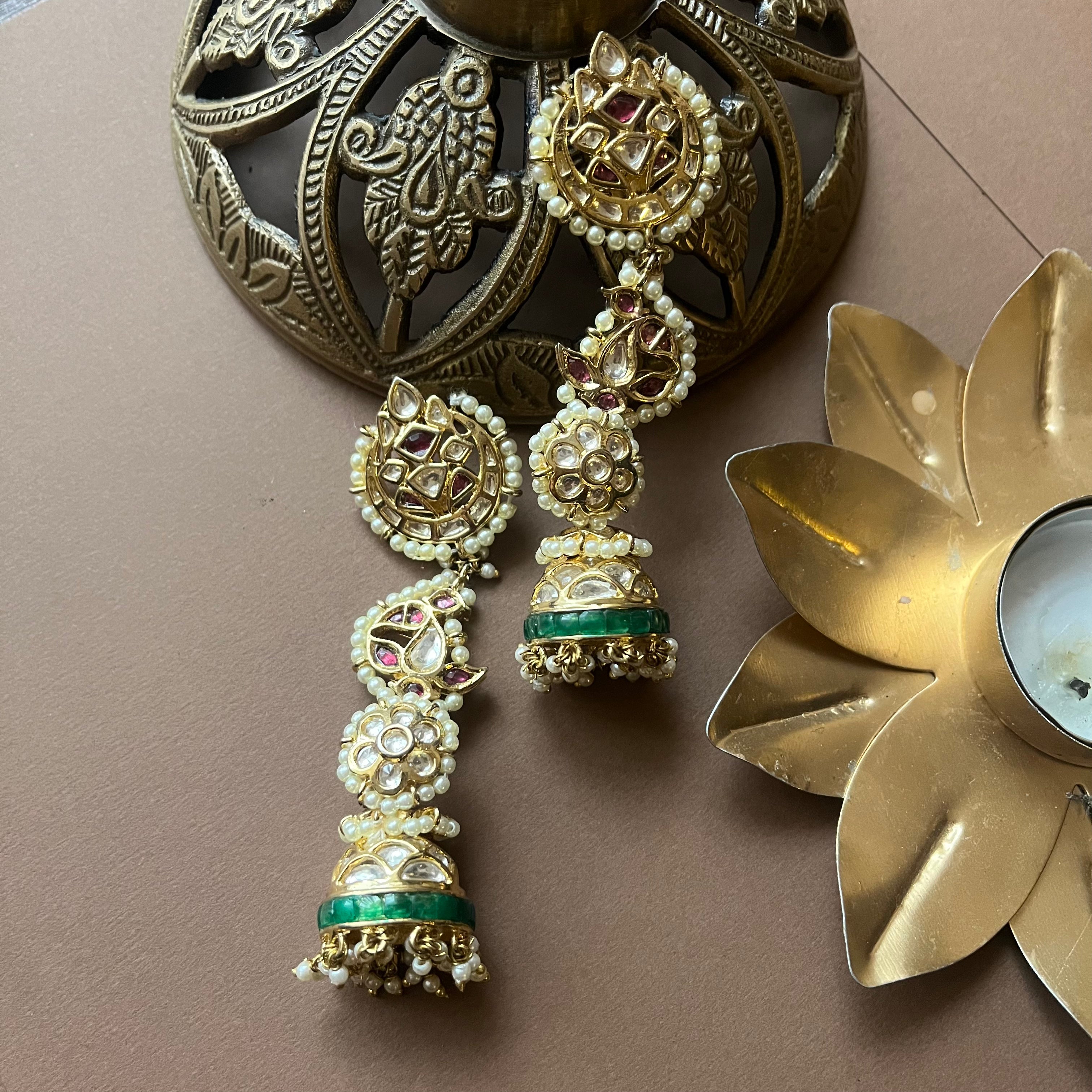 Kavisha Kundan jhumka earrings in gold, pink, and green placed elegantly on antique brass decorative background.